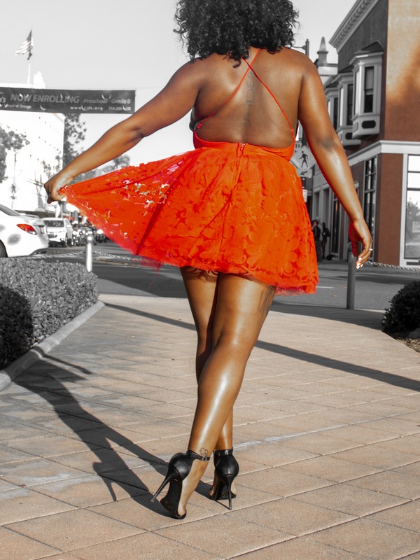 elegant black curvy lady viewed from the back with curly black hair, a lace short red dress and black high heels decollete shoes walking in the street