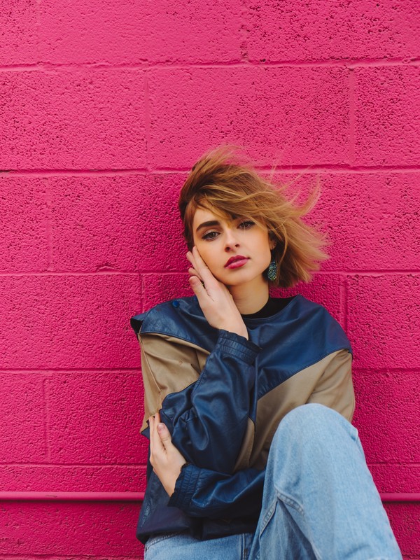 stylish redhead girl with blue eyes, hot pink lipstick, a blue and beige rain jacket and blue jeans sitting on the floor against a hot pink wall with a pensive expression on her face
