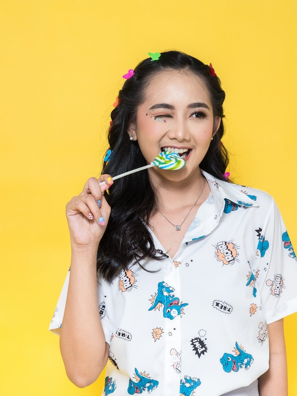 stylish asian woman with many colorful hair clips on her hair eating a lollipop while winking and wearing a white shirt with funny blue t-rex prints and a yellow background