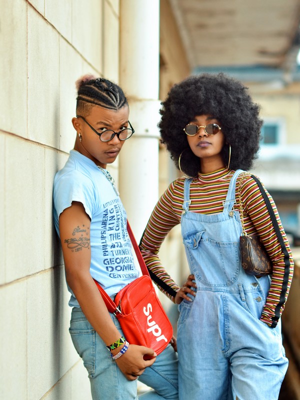 two black people, potentially non-binary both wearing a casual outfit the one on the left has cornrows a light blue tshirt a red cross body bag and blue jeans and is leaning against a white wall, the one on the right has afro hair, hoop earrings, sunglasses, jeans overall with a yellow and red horizontal stripes top and a mini louis vuitton bag and she is standing next to the other one