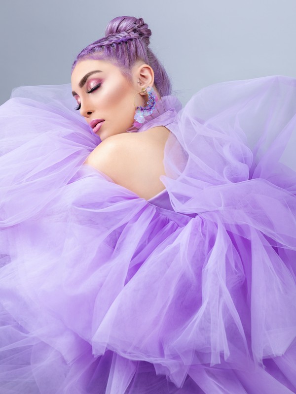 a white woman with lilac hair tied into a up hairdo with a braid, lilac long earrings, pink eyeshadow and pink lipstick, her eyes are closed and she is wearing a big lilac tulle dress