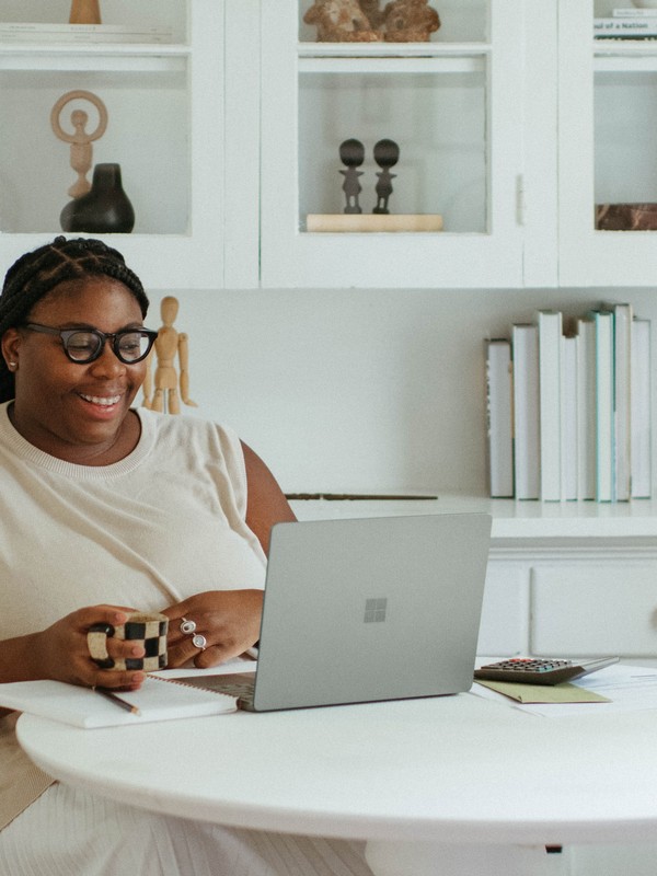 a curvy black woman smiling while looking at something on her silver laptop while wearing black glasses, cornrows on her hair a white tshirt and holding a coffee mug with a big white cabinet in the background