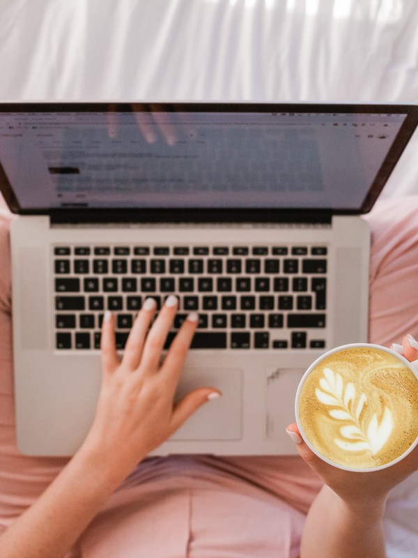 a macbook computer on top of a woman's lap where you can see her pink pants and pink shirt while one of her hands is typing on it and the other one is holding a flat white with a leaf coffee art