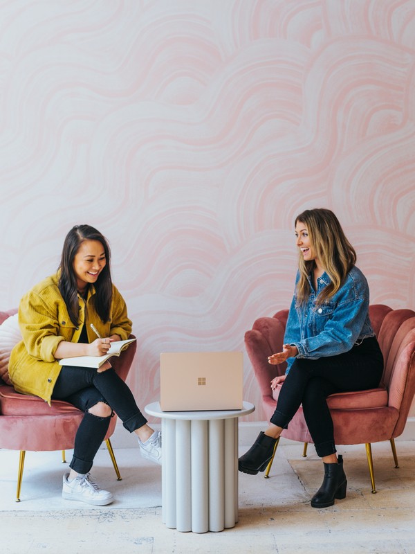 on the left there is a black haired woman wearing a yellow jacket black pants and white runners while holding a notebook and a pencil smiling and sitting on a pink armchair talking to the woman on the right who has dyed dark blonde hair and wears a blue jeans jacket black pants and black shoes while looking at the computer and sitting on another pink armchair with a pink background
